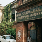 Vintage-style photograph of the red brick entrance of the Indian Psychoanalytical Society in Kolkata, featuring a weathered signboard with the text "Indian Psychoanalytical Society Estd. 1922" in English and Bengali.
