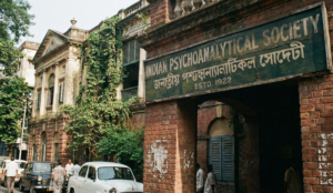 Vintage-style photograph of the red brick entrance of the Indian Psychoanalytical Society in Kolkata, featuring a weathered signboard with the text "Indian Psychoanalytical Society Estd. 1922" in English and Bengali.