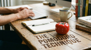 A person's hands typing on a keyboard at a wooden desk with a notebook, pen, coffee mug, a stack of psychology books, and a red tomato-shaped timer. The words "THE POMODORO TECHNIQUE" are superimposed on the desk surface.