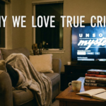 A woman curled on a sofa under a blanket at night, watching the 'Unsolved Mysteries' documentary series on a television screen. In the foreground, a stack of books labeled "TRUE CRIME" rests on a wooden coffee table next to a mug and an open journal. The text overlay reads "WHY WE LOVE TRUE CRIME."