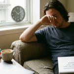 A tired man sits on a couch with his hand over his eyes, next to a stack of self-help books and a notebook that reads "Burnout Recovery: Why a Weekend Off Isn't Enough."