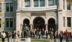 A diverse group of professionals and students walking outside a classic stone academic building with a large banner reading American Psychological Association.