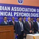 A formal conference stage featuring a large blue banner reading "Indian Association of Clinical Psychologists" with dignitaries and professionals standing in formal attire.