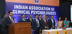 A formal conference stage featuring a large blue banner reading "Indian Association of Clinical Psychologists" with dignitaries and professionals standing in formal attire.