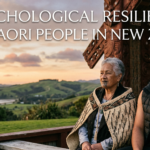 Photograph of an elder Maori woman with a moko kauae and a young Maori man with arm ta moko, sitting together on a bench. They are outside a carved wharenui (meeting house) with a coastal New Zealand landscape at sunset. White text at the top reads: 'THE PSYCHOLOGICAL RESILIENCE OF THE MAORI PEOPLE IN NEW ZEALAND'.